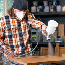Man wearing mask using Katana Plus HVLP Spray Gun to spray wood surface with smooth finish in workshop setting